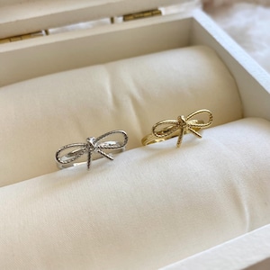 Op de afbeelding: Twee ringen, één zilverkleurig en één goudkleurig, elk met een strikdesign. De ringen worden gepresenteerd in een witte sieradendoos met een satijnen voering. De strikdetails zijn delicaat.