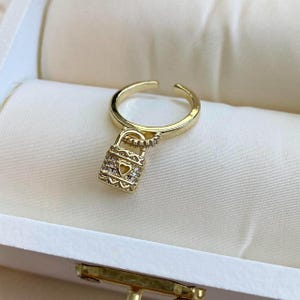 May include: Gold ring with a padlock charm adorned with clear cubic zirconia stones. The padlock has a heart-shaped cutout.