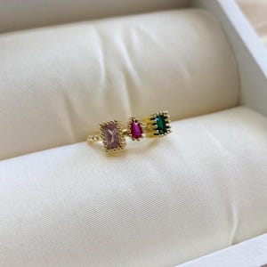 May include: Gold ring with three colorful gemstones. The ring features a pink rectangular stone, a red teardrop-shaped stone, and a green rectangular stone.
