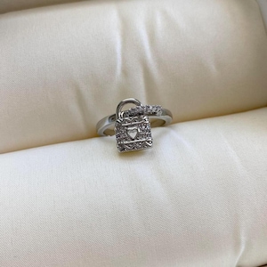 May include: A silver ring with a padlock design. The padlock is encrusted with small clear stones and has a heart-shaped opening. The ring band is also decorated with small clear stones.