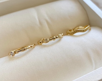 May include: Four gold rings displayed on a white satin surface inside a jewelry box. The rings have different designs, including a chain link, beaded, and wavy style. One ring features a small gemstone. The rings are in a jewelry box.