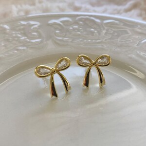 May include: A pair of gold-colored bow-shaped stud earrings. Each earring features a clear, teardrop-shaped stone at the center of the bow. The earrings are displayed on a white, decorative plate.