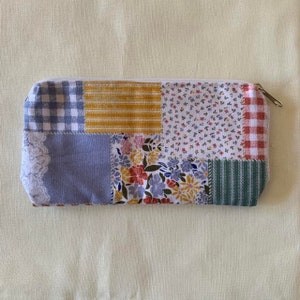 May include: A patchwork fabric zippered pouch with a variety of patterns, including gingham, floral, and stripes in shades of blue, yellow, red, and green.