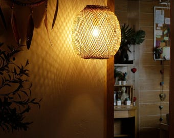 以下が含まれることがあります： 丸い籐製のペンダントライトが天井から吊り下げられています。ライトは点灯しており、後ろの壁に温かい光を投げかけています。