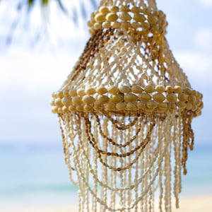 Puede incluir: Una lámpara de araña decorativa hecha de conchas marinas, con un esquema de color blanco y beige. La lámpara de araña está colgada de una rama de árbol, con un cielo azul y el océano en el fondo.