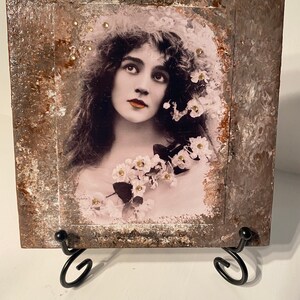 Puede incluir: Una fotografía vintage de una mujer con el pelo largo y oscuro, vestida con un vestido blanco con flores, enmarcada en un marco de madera marrón rústico con detalles dorados.