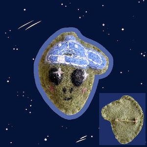 May include: A green felt alien wearing a blue and white felt hat with a star on its forehead. The alien has black eyes and a smiling mouth. The back of the pin is shown with a gold pin back.