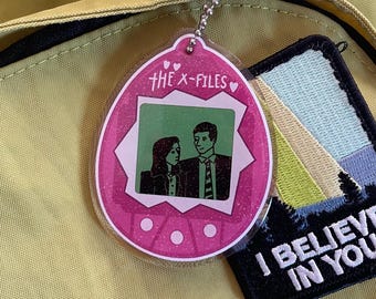 Może przedstawiać: Różowy plastikowy brelok w kształcie Tamagotchi z napisem "THE X-FILES" i czarno-białym obrazkiem dwóch osób w środku. Brelok jest przymocowany do srebrnego łańcuszka.