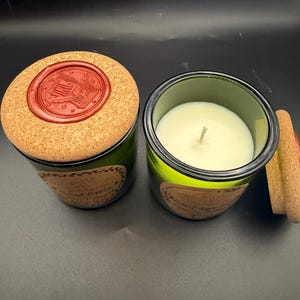 May include: Two green glass candle jars with white wax and single wicks. Each jar features a tan paper label and a round cork lid topped with a red wax seal embossed with an open book design. One lid is placed on a jar, while the other sits beside it.