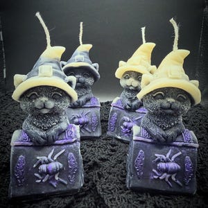 May include: Four black cat candles, each wearing a cream-colored witch hat. The cats are emerging from dark purple rectangular bases decorated with raised spider motifs. Each candle features a single vertical wick extending from the hat top.