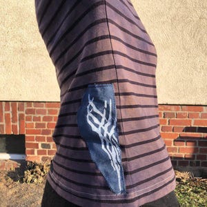 Op de afbeelding: Een gestreept paars en zwart t-shirt met een blauwe patch en wit ontwerp. Het t-shirt heeft horizontale strepen en een opvallende patch aan de zijkant genaaid. De stijl is casual en artistiek.