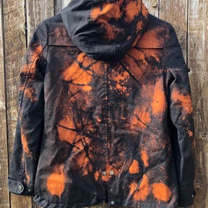 May include: Black hooded jacket with an orange bleach-dye design. The jacket has a button-up front, a hood, and pockets. The orange dye creates a unique, abstract pattern across the jacket's surface.