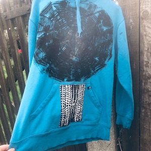 Könnte beinhalten: Ein türkisfarbener Kapuzenpullover mit einem großen schwarzen kreisförmigen Design auf der Vorderseite. Der Hoodie hat eine Kängurutasche mit einem schwarz-weißen Muster. Er hat eine Kapuze mit Kordelzug und hängt an einem Metallbügel.