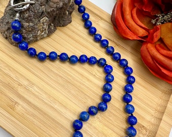 May include: A long necklace made of dark blue lapis lazuli beads with a silver toggle clasp. The necklace is lying on a wooden surface.