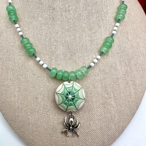 Puede incluir: Un collar de cuentas verdes con un colgante de telaraña y un dije de araña plateada. El collar presenta cuentas verdes y blancas. El colgante es blanco con un diseño de telaraña verde.