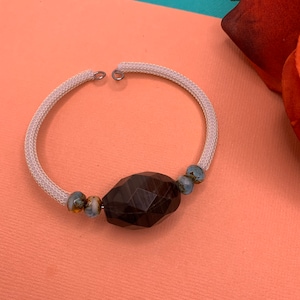 Puede incluir: Un brazalete elástico blanco con una gran cuenta facetada de color marrón oscuro y cuentas más pequeñas de color azul y marrón.