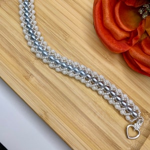 Peut inclure: Un bracelet en perles argentées et blanches avec un fermoir en forme de cœur. Le bracelet est fait de petites perles rondes dans un motif tissé.