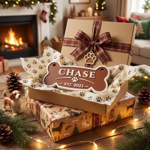 May include: A gift box with a brown plaid ribbon and a paw print charm. Inside, a bone-shaped sign with the name "CHASE" and "EST. 2023". The box sits atop a dog-themed wrapping paper, with a small dog figurine nearby.
