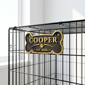 May include: Black and gold dog bone-shaped sign with the name "COOPER" in gold lettering. The sign also includes a paw print and the text "EST. 2024". The sign is attached to a black metal dog crate.