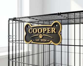 May include: Black and gold dog bone-shaped sign with the name "COOPER" in gold lettering. The sign also includes a paw print and the text "EST. 2024". The sign is attached to a black metal dog crate.