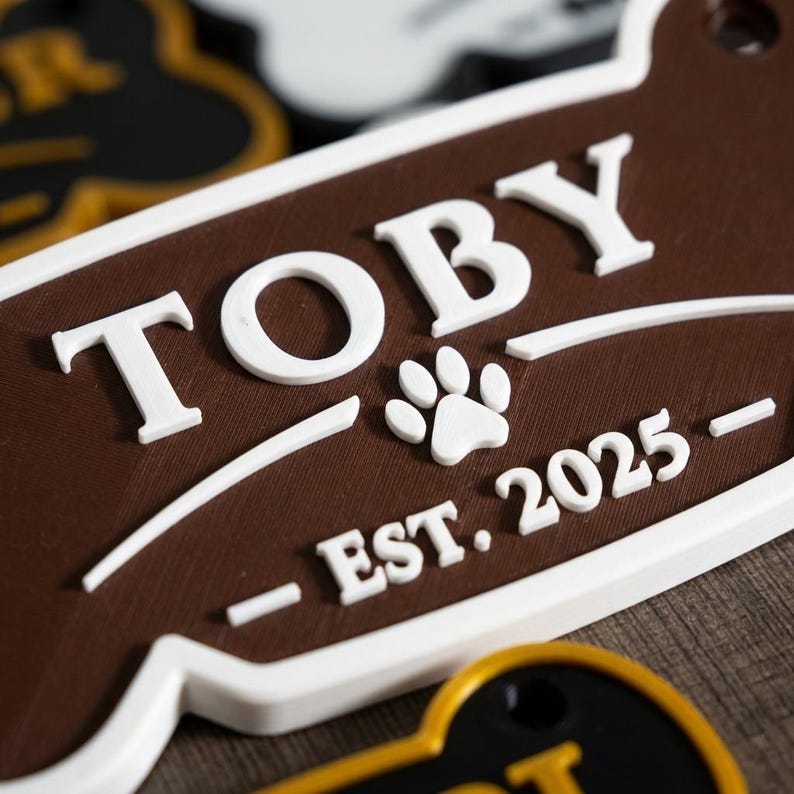 May include: A brown and white pet tag with the name "TOBY" in white, along with a paw print and the text "EST. 2025". The tag has a rounded shape with a keyhole for attachment.