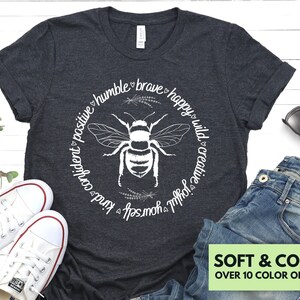 May include: A dark gray t-shirt with a white bee graphic and the text "humble brave happy wild creative joyful kind confident positive" in a circle around the bee.