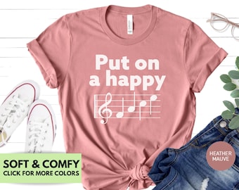 Może przedstawiać: Różowy T-shirt z napisem "Put on a happy" i pięciolinią z nutami. Koszulka jest miękka i wygodna i dostępna w innych kolorach.