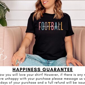 Op de afbeelding: Een zwart T-shirt met de tekst "FOOTBALL mama" in regenboogkleuren. De tekst is in een speels lettertype met een hartje. Het shirt wordt gedragen door iemand die op een roze bank zit.