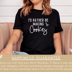 May include: A black t-shirt with the text "I'd rather be making cookies" in white lettering. The shirt is being worn by a person sitting on a pink couch. The text is surrounded by small white hearts.