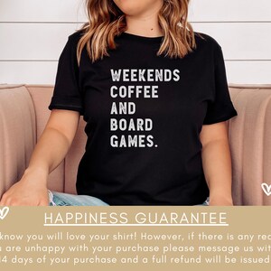 May include: A black t-shirt with white text that reads "Weekends Coffee And Board Games." The shirt is being worn by a person sitting on a pink couch. The background is a white wall with a green plant.