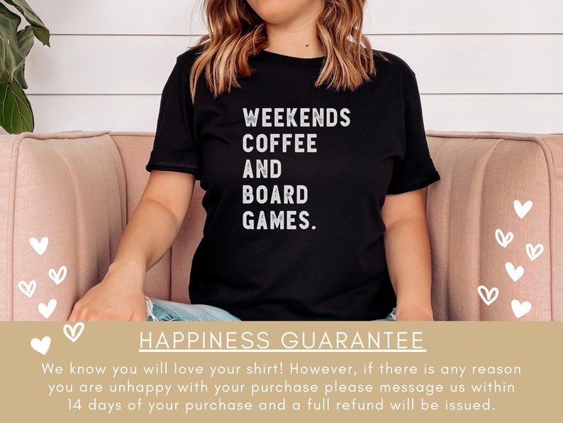 May include: A black t-shirt with white text that reads "Weekends Coffee And Board Games." The shirt is being worn by a person sitting on a pink couch. The background is a white wall with a green plant.
