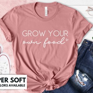 May include: A dusty rose colored t-shirt with the text "GROW YOUR own food" in white lettering with a small heart symbol. The t-shirt is made of a soft material and is available in a variety of colors.