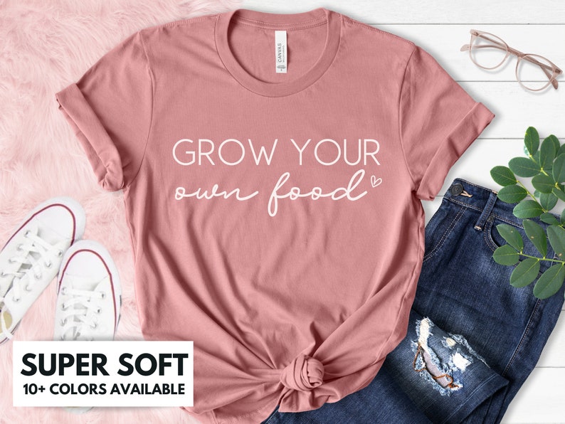 May include: A dusty rose colored t-shirt with the text "GROW YOUR own food" in white lettering with a small heart symbol. The t-shirt is made of a soft material and is available in a variety of colors.