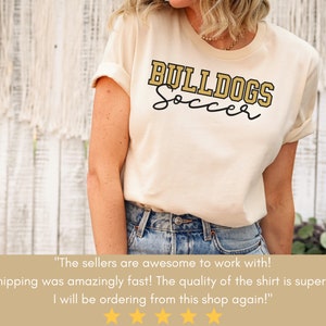 May include: A beige t-shirt with the text "BULLDOGS Soccer" in gold lettering. The shirt is being worn by a person. The text "The sellers are awesome to work with! Shipping was amazingly fast! The quality of the shirt is superb! I will be ordering from this shop again!" is displayed below the image.