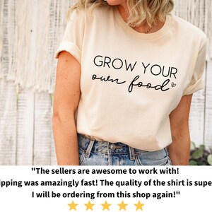May include: A light beige t-shirt with the text "GROW YOUR own food" in black lettering. The shirt is being worn by a person with their arms at their sides. The background is a white wooden wall.