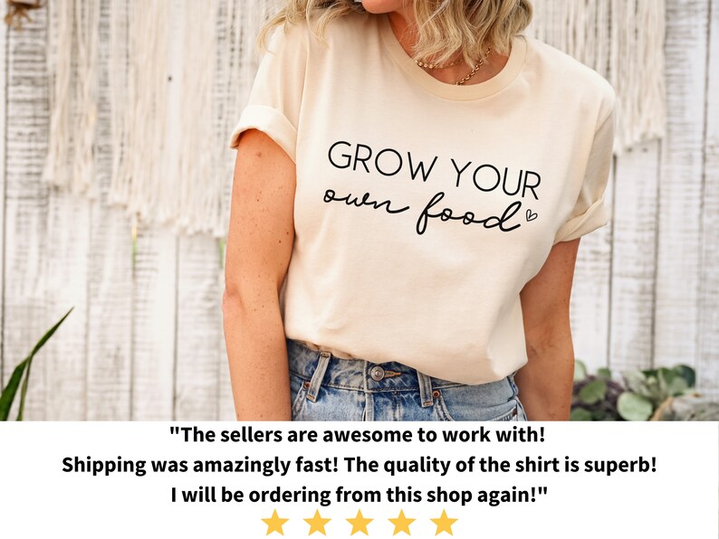 May include: A light beige t-shirt with the text "GROW YOUR own food" in black lettering. The shirt is being worn by a person with their arms at their sides. The background is a white wooden wall.
