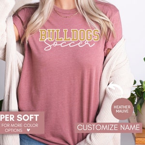 May include: A heather mauve t-shirt with the text "BULLDOGS Soccer" in gold lettering. The shirt is super soft and available in other colors. Customize the name on the shirt.