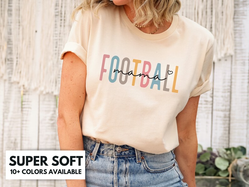 Op de afbeelding: Een cr&egrave;mekleurig T-shirt met de tekst "FOOTBALL mama" in regenboogkleuren. De tekst is in een gestileerd lettertype met een hartje na "mama".