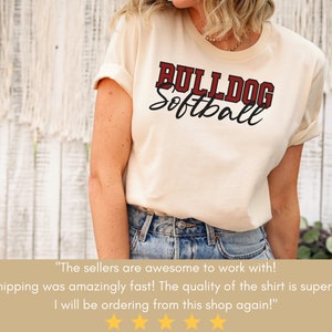 May include: A woman wearing a cream colored t-shirt with the text "BULLDOG Softball" in red and black lettering. The shirt is a casual style with rolled up sleeves. The woman is wearing blue jeans.