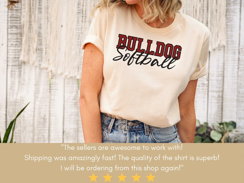 May include: A woman wearing a cream colored t-shirt with the text "BULLDOG Softball" in red and black lettering. The shirt is a casual style with rolled up sleeves. The woman is wearing blue jeans.