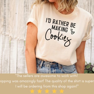 May include: A beige t-shirt with the text "I'd rather be making cookies" in black lettering. The shirt has a rolled up sleeve and is being worn by a person.