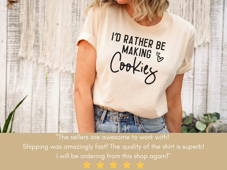 May include: A beige t-shirt with the text "I'd rather be making cookies" in black lettering. The shirt has a rolled up sleeve and is being worn by a person.