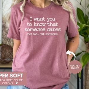 以下が含まれることがあります： 「I want you to know that someone cares - not me, but someone -」の文字が入った、ヘザーモーヴ色のTシャツ。「Super Soft」と「Click for more color options」の文字もプリントされています。