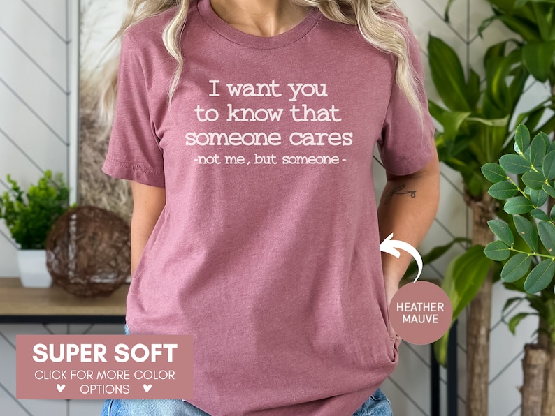 以下が含まれることがあります： 「I want you to know that someone cares - not me, but someone -」の文字が入った、ヘザーモーヴ色のTシャツ。「Super Soft」と「Click for more color options」の文字もプリントされています。