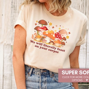 Op de afbeelding: Een beige T-shirt met een afbeelding van paddenstoelen en de tekst "We will literally feast on your corpse". Het shirt is super zacht en verkrijgbaar in verschillende kleuren.