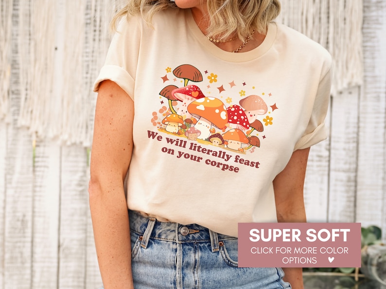 Op de afbeelding: Een beige T-shirt met een afbeelding van paddenstoelen en de tekst "We will literally feast on your corpse". Het shirt is super zacht en verkrijgbaar in verschillende kleuren.