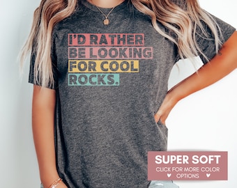 Könnte beinhalten: Ein heidefarbenes T-Shirt mit dem Text "I'D RATHER BE LOOKING FOR COOL ROCKS." in bunten Blockbuchstaben.