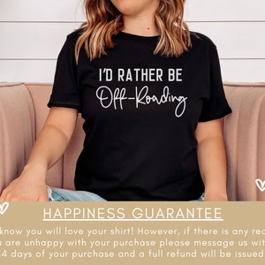 May include: A black t-shirt with white text that reads "I'd rather be Off-Roading". The shirt is being worn by a person sitting on a pink couch. The background is a white wall with a green plant.