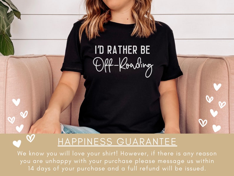 May include: A black t-shirt with white text that reads "I'd rather be Off-Roading". The shirt is being worn by a person sitting on a pink couch. The background is a white wall with a green plant.
