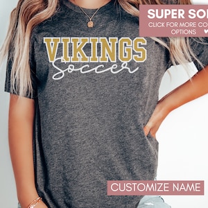 May include: A gray t-shirt with the text "VIKINGS Soccer" in gold and white lettering. The text "SUPER SOFT" and "CLICK FOR MORE COLOR OPTIONS" is displayed in pink at the top of the image. The text "CUSTOMIZE NAME" is displayed in pink at the bottom of the image.
